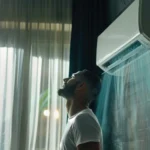 A man standing in front of a sleek wall-mounted split AC unit enjoying the cool breeze, illustrating the concept of how does air conditioning remove humidity and cool indoor air.