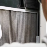 A technician wearing white gloves slides a dusty pleated air filter into a home HVAC system, illustrating what does merv mean on furnace filters.