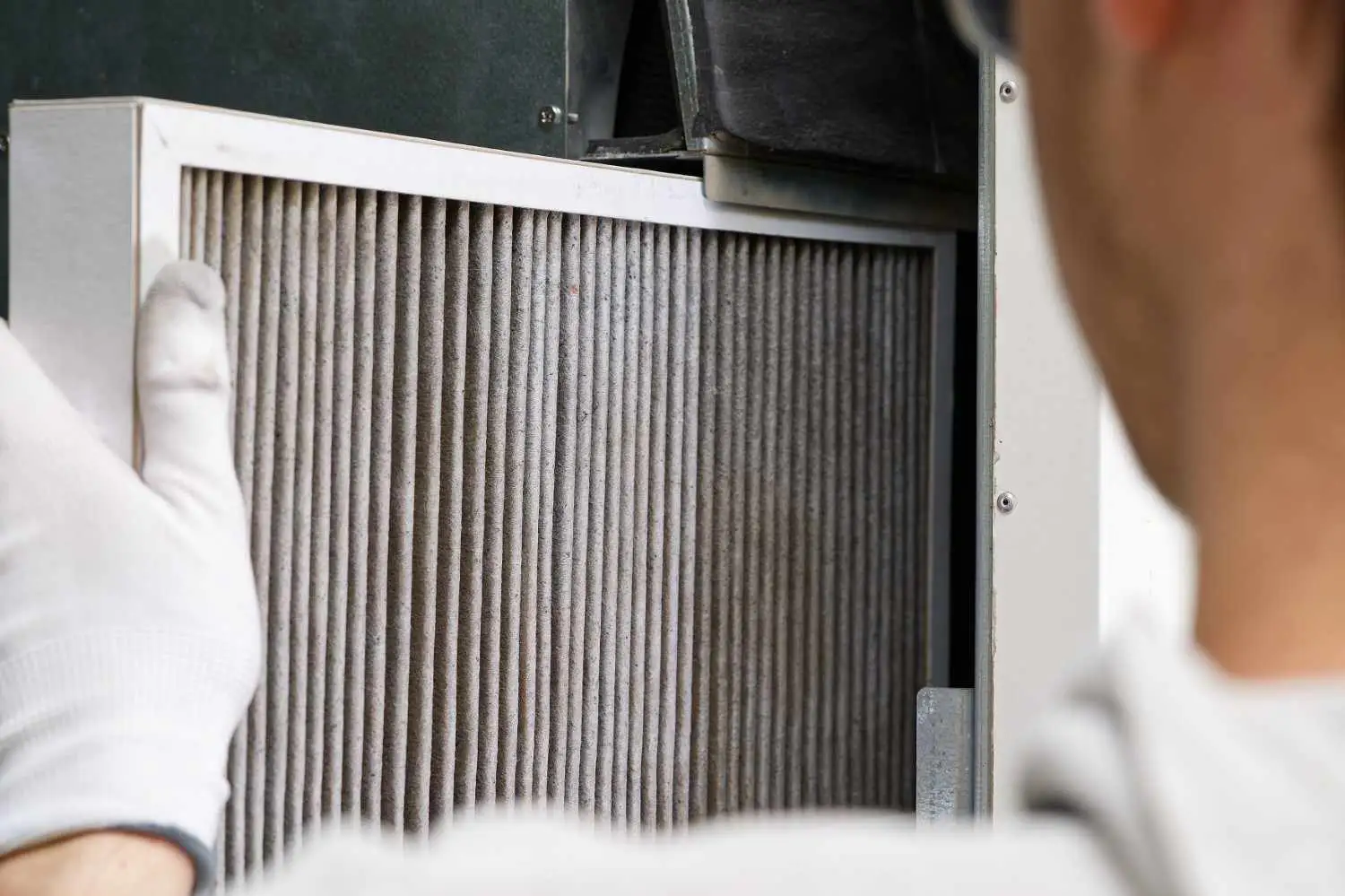 A technician wearing white gloves slides a dusty pleated air filter into a home HVAC system, illustrating what does merv mean on furnace filters.