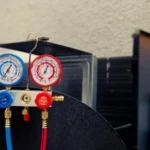 A close-up of blue and red refrigerant manifold gauges hooked up to an outdoor air conditioning unit, illustrating how technicians measure performance to explain what does SEER mean in HVAC systems.