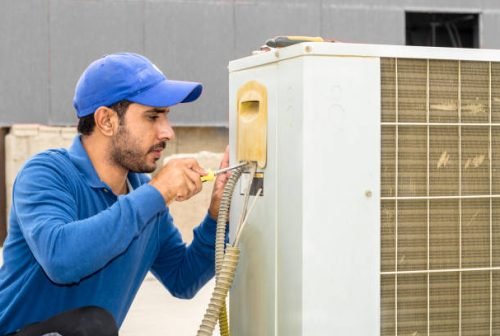 a professional electrician man is fixing a heavy duty unit of central air conditioning system by his tools on the roof top and wearing grey color of uniform and white cap